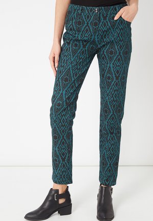Mujer con pantalones de estampado geométrico en tonos verde azulado y negro y botines negros, de pie con una mano en el bolsillo contra un fondo blanco.