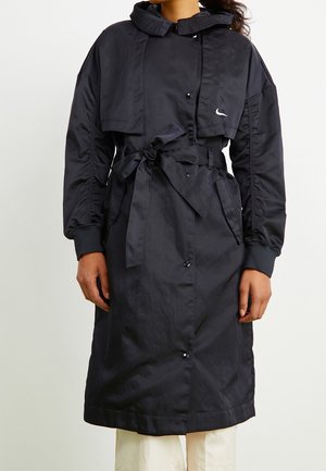 Vrouw draagt een lange zwarte jas met ceintuur, hoge kraag en een klein wit logo op de borst, gecombineerd met lichtgekleurde broek.