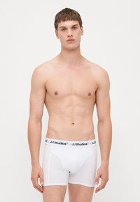 Weiße Boxer-Briefs mit schwarzem "JJ Studios"-Bund, glatte Textur, taillierte Passform und einfarbig. Das Modell steht vor neutralem Hintergrund.