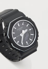 Zegarek G-Shock z czarnym paskiem z żywicy, ośmiokątną kopertą, analogowym i cyfrowym wyświetlaczem, białymi oznaczeniami godzin oraz srebrnymi elementami ozdobnymi. Wodoodporny.
