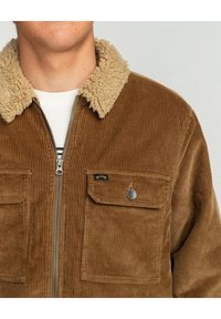 Ruskea corduroy-takki, jossa on vaalea keinoturkiskaulus, etujakaja ja kaksi rintataskua. Sisältää pystysuoran ribbitekstuurin ja logotagin.