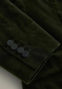 Rękaw ciemnozielonej aksamitnej kurtki z czterema teksturowanymi czarnymi guzikami oraz delikatnymi detalami haftu. Gładka, miękka tekstura tkaniny.
