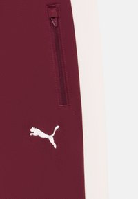 Bordowe dresy sportowe z białą wstawką, z kieszenią na zamek błyskawiczny i białym haftowanym logo skaczącego kota po lewej stronie.