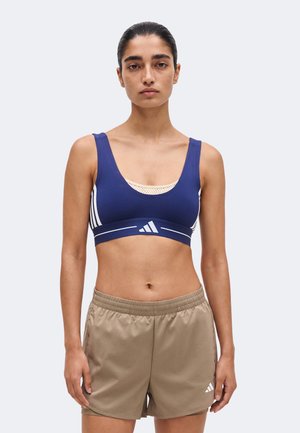Kobieta stojąca przodem, ubrana w granatowy sportowy biustonosz i beżowe szorty sportowe z logo Adidas na jednolitym tle.