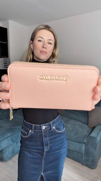 Šviesiai rožinė odinė piniginė su tekstūruotu paviršiumi, auksiniu „VALENTINO“ logotipu, užtrauktuko uždarymu ir auksinėmis detalėmis. Matmenys nenurodyti.