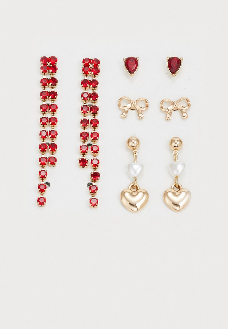 Drie paar oorbellen: rode stenen studs, gouden hart-drops met witte parels en gouden strik-studs. Allemaal met glanzende metallic afwerkingen.