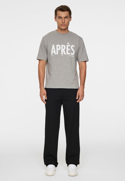Mężczyzna stojący, ubrany w szarą koszulkę z krótkim rękawem z napisem "APRÈS", czarne spodnie i białe trampki na jednolitym białym tle.