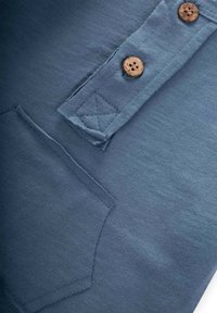 La tela de algodón azul muestra un bolsillo de parche y dos botones de madera en una tapeta con un acabado texturizado y detalles cosidos.