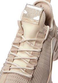 Beige sneaker with knit upper and reinforced heel. Features textured patterns, flat laces, and a shiny, studded accents on the tongue.