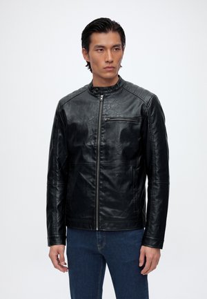 Homme portant une veste en cuir noire zippée avec une poche zippée sur la poitrine et un jean bleu, debout devant un fond blanc uni.
