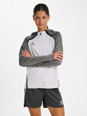 Kobieta stoi z założonymi ramionami, ubrana w jasnoszarą bluzę z kapturem i zamkiem oraz ciemnoszare szorty z logotypami marek sportowych.