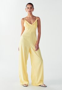 Mujer vestida con un mono sin mangas de pierna ancha amarillo y sandalias blancas de punta abierta, de pie frente a un fondo claro y liso.