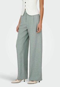 Femme portant un pantalon large gris clair, un blazer court blanc et des chaussures beige à talons bas pointues, debout les mains relâchées.