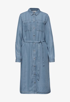 Femme portant une robe en jean bleu clair avec les manches retroussées et une ceinture nouée, la main posée dans la poche latérale.