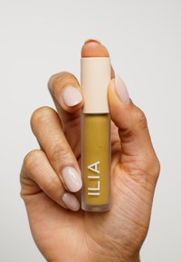 Prodotto per le labbra ILIA tenuto in mano, con un contenitore trasparente giallo e un cappuccio beige, che mostra il nome del marchio in lettere bianche.