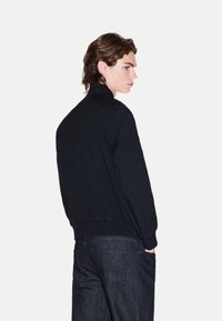 Giubbotto bomber nero in tessuto texture, con colletto e polsini a costine, caratterizzato da un design aderente e un delicato motivo trapuntato, abbinato a jeans scuri.