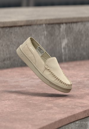 Mocassin slip-on beige en daim avec un panneau latéral matelassé, des coutures style mocassin et une semelle en caoutchouc, flottant au-dessus d'une surface en pierre.