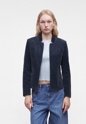Jeune femme aux cheveux blonds portant une veste en velours côtelé bleu marine foncé par-dessus une chemise courte bleu clair et un jean bleu, debout sur un fond blanc.