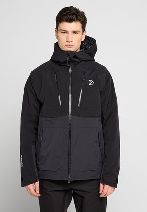 Mann trägt eine schwarze und dunkelgraue Kapuzenjacke mit Frontreißverschluss und Reißverschlusstaschen, steht vor einem schlichten hellen Hintergrund.