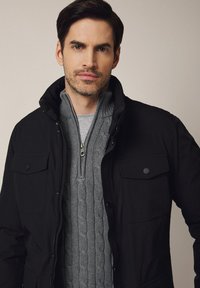 Czarna kurtka z wysokim kołnierzem i zapinanymi kieszeniami, założona na szary sweter w warkocze. Gładka faktura, zamek błyskawiczny z przodu.