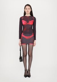 Black sheer dress with red lace accents, long sleeves, and a fitted shape. The model holds a small black handbag, wearing black tights and heels.