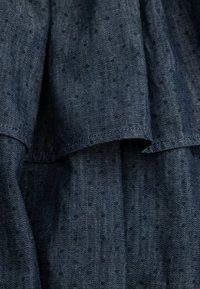 Denim w ciemnoniebieskim odcieniu z teksturowaną powierzchnią, na której znajdują się małe, równomiernie rozmieszczone kropki. Zawiera warstwowe falbany na krawędzi.