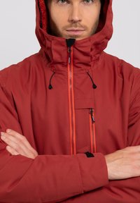 Chaqueta impermeable roja con un cuello alto y capucha ajustable. Presenta cremalleras en contraste y un bolsillo en el pecho. Material texturizado, diseño entallado.