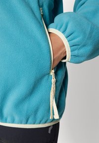 La veste en polaire turquoise présente un col montant, des poignets élastiqués et une ceinture contrastante beige avec un détail de poche zippée sur le côté.