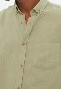 Camisa abotonada verde claro hecha de tela texturizada, con un bolsillo en el pecho y botones beige. Detalles del cuello y el tapeta son visibles.