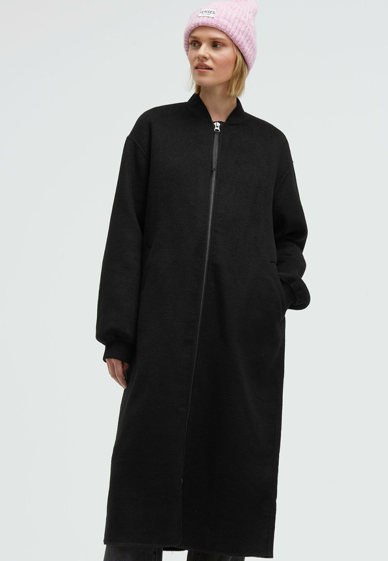 Long black fleece coat with a high collar and front zipper, featuring oversized sleeves and side pockets. Model wears a pink knitted beanie.