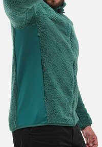 Mann in einer strukturierten Fleecejacke in tealgrün mit seitlichem Reißverschluss und schwarzen Hosen, Arm erhoben, oberer Gesichtsauschnitt sichtbar.