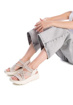 Sandalias de tiras beiges con una superficie texturada, punta abierta y suela blanca gruesa, que se llevan con pantalones grises cortos.