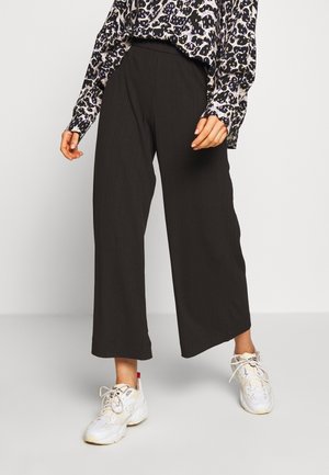 Vrouw draagt wijde zwarte cropped broek, witte sneakers en een langarmige top met luipaardprint, staand op een lichtgrijze vloer.