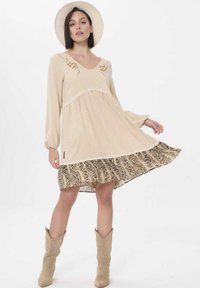 Beige jurk met lange mouwen, V-hals en kantafwerking; beschikt over een gestructureerde slangenprint met een rucheszoom. Gecombineerd met beige cowboylaarzen.