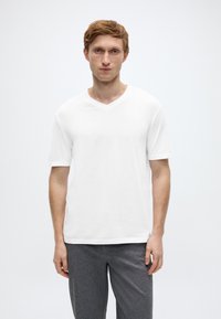 Λευκή μπλούζα με V λαιμόκοψη από ελαφρύ ύφασμα με κοντά μανίκια, συνδυασμένη με γκρι παντελόνι. Απλός σχεδιασμός χωρίς σχέδια ή εμφανή λογότυπα.