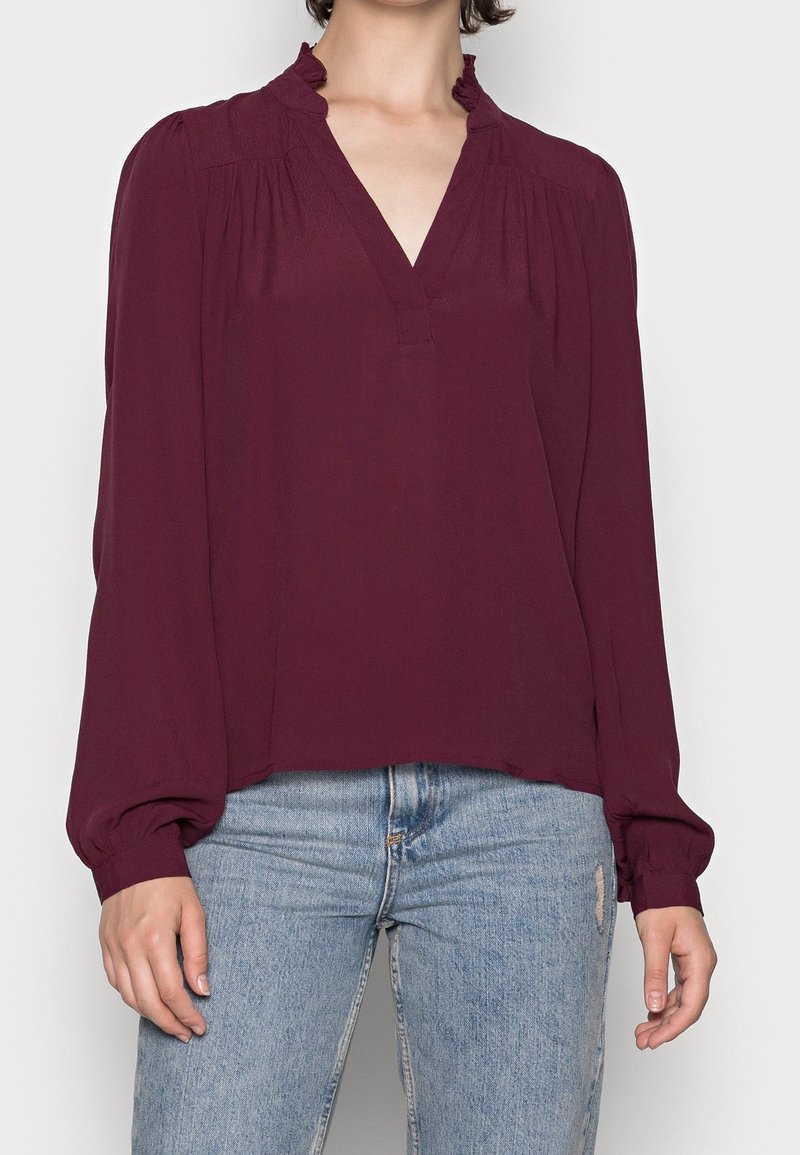 Femme portant un chemisier bordeaux à manches longues avec un col en V à volants et un jean bleu clair légèrement usé sur la cuisse droite.