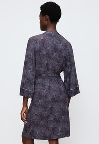 Donna che indossa un vestito da ginocchio a maniche larghe di un viola scuro con motivo floreale, vista da dietro su uno sfondo bianco.