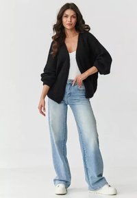 Czarny dzianinowy kardigan z szerokimi rękawami, w połączeniu z jasnoniebieskimi spodniami o wysokim stanie i białymi sneakersami, noszony przez modelkę.