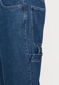 Tummansiniset farkkukankaiset cargo-housut, joissa on näkyvät saumat, sivutaskut ja teksturoitu pinta. Ominaista rento istuvuus ja kestävä materiaali.