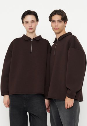 Deux jeunes portent des pulls surdimensionnés marron foncé à fermeture éclair quart de zip avec col et pantalons foncés, debout sur un fond blanc uni.