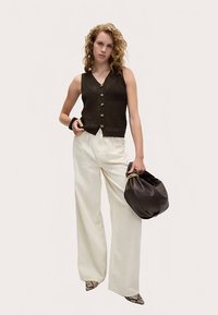 Top nero senza maniche in crochet con bottoni frontali, abbinato a pantaloni bianchi larghi. La modella tiene una borsa nera, che esalta il contrasto dell'outfit.