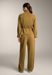 Compleu sport de culoare verde măsliniu, cu guler înalt, talie strânsă și pantaloni cu picioare largi. Material neted cu detalii vizibile ale cusăturii.