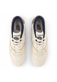 White sneakers with a textured upper, dark blue accents, beige logo, and flat laces. Features a padded collar and perforated toe. Model 550.