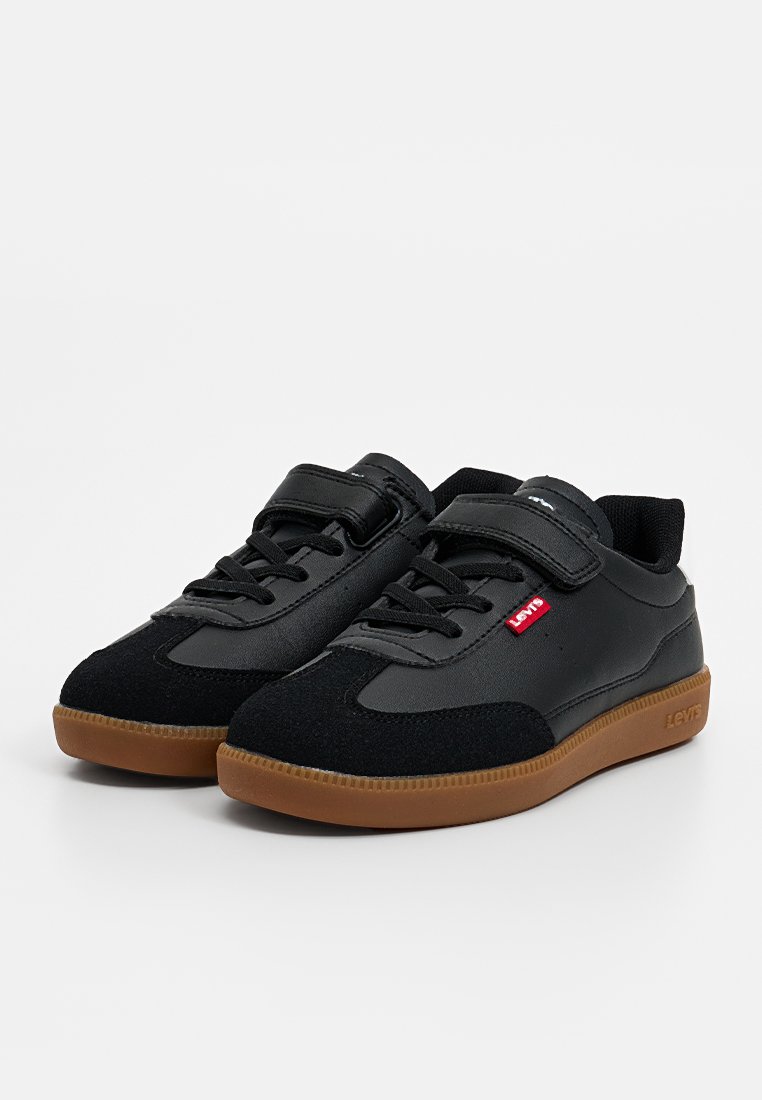 Zwarte sportschoenen van een combinatie van gladde en gestructureerde materialen, met veters en klittenbandsluitingen, en een bruine rubberen zool. Rode logodetail.