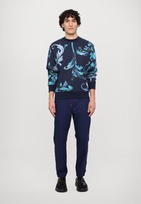 Granatowy sweter z turkusowym kwiatowym wzorem, z prążkowanymi mankietami i dołem, w połączeniu z granatowymi spodniami i czarnymi butami. Gładka faktura, casualowy krój.