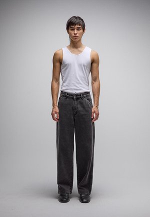 Noor mees seisab halli tausta ees, kandes valget varrukateta särk, lahtisi musti teksapükse, musti kingi ja hõbedasi aksessuaare.