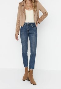 Chaqueta de moto de ante color tostado, top ajustado crema, jeans ajustados azules y botas de tobillo tostadas con un acabado texturizado y tacón cuadrado.