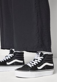 Zwarte high-top sneakers met witte accenten, textuur van suede materiaal, witte veters en een klassiek Vans ontwerp. Donkere, iets gekreukte stof in de buurt.
