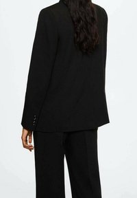 Blazer noir sur mesure à manches longues, tissu lisse et poignets boutonnés, associé à un pantalon large assorti. Vue de dos affichée.