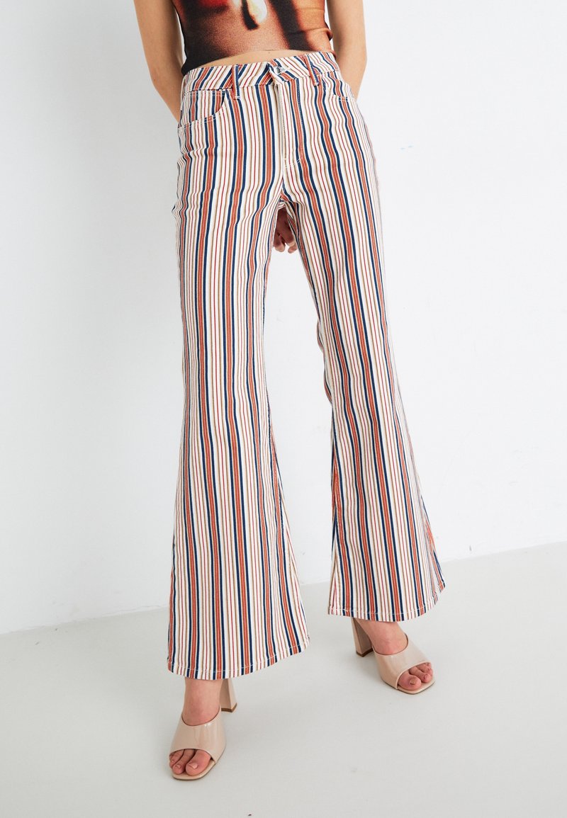 Personne portant un pantalon évasé taille haute à rayures verticales multicolores et des chaussures à talons ouverts beiges, debout sur un fond blanc uni.